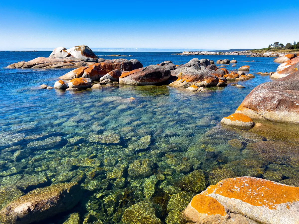The water at the edge of the bay is crystal clear, revealing the rocky floor beneath the bright orange boulders.