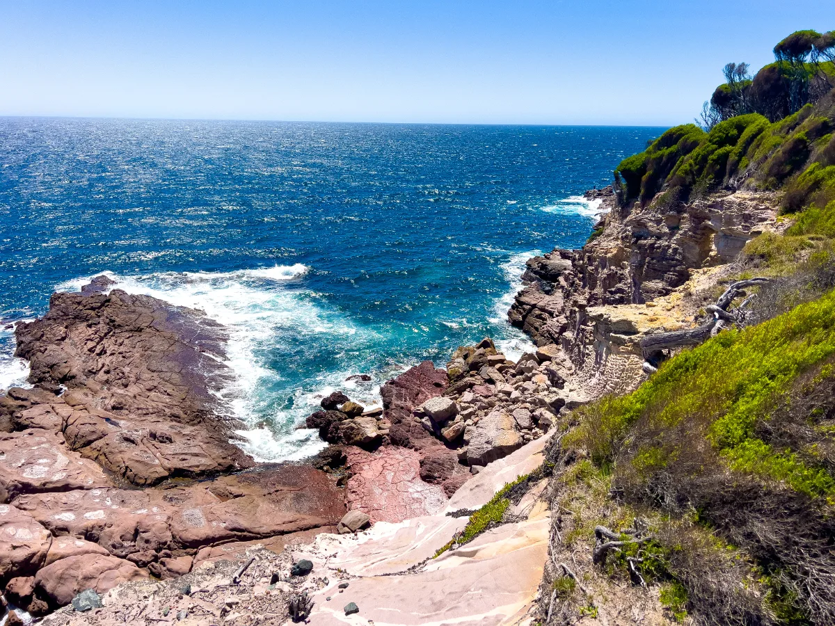 Rugged, red-hued rock formations meet the churning surf along the Tura Beach coastline.