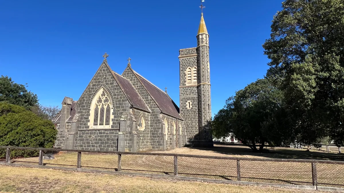 Birregurra's stone church, stoic and enduring against a backdrop of blue sky and verdant trees.